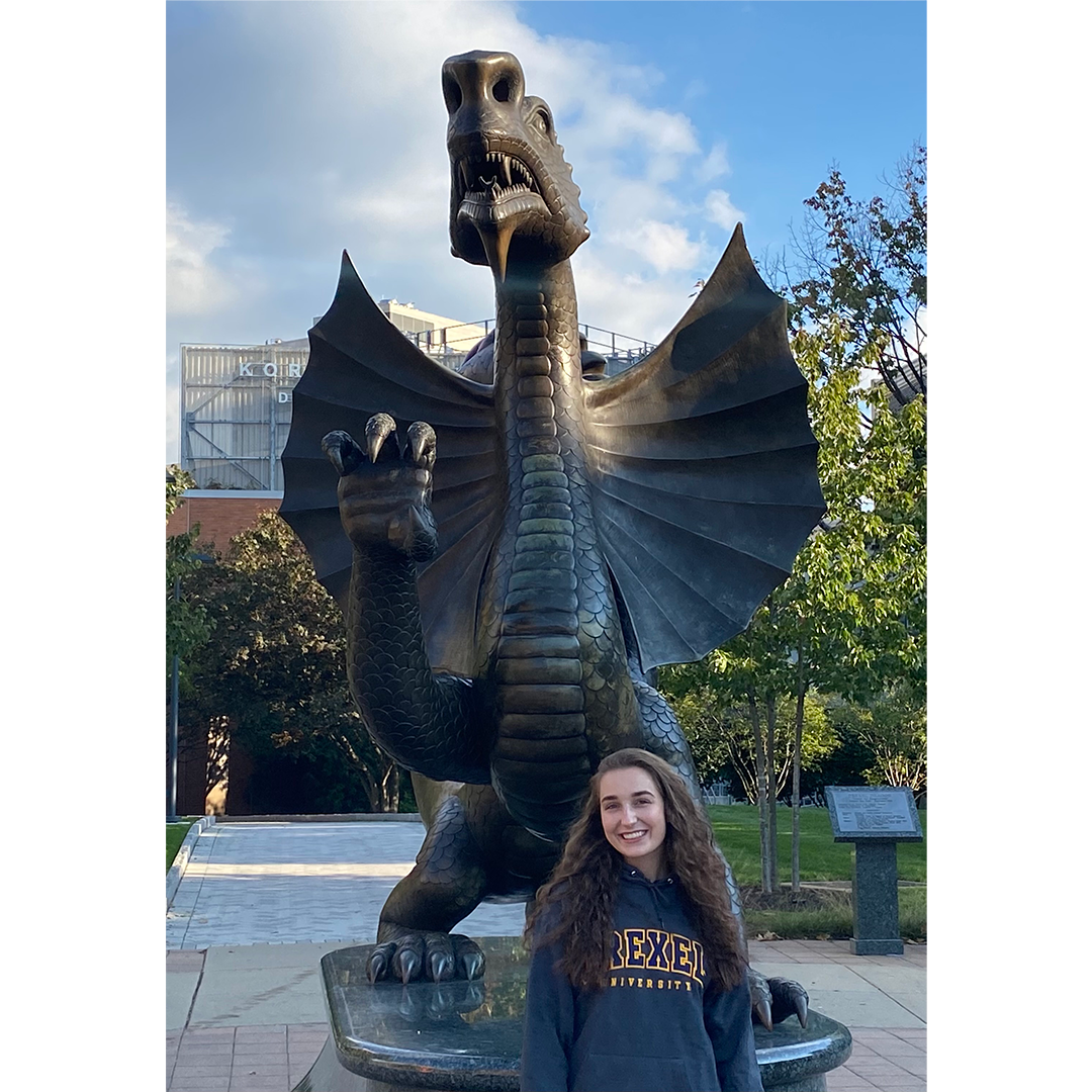Grace Schuler sits in front of Mario the Dragon statue on Drexel main campus, Grace is wearing a Drexel sweatshirt 
